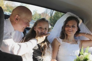 Narzeczeni jadący samochodem na ceremonię. Pan młody bawi się z siedzącą pomiędzy nimi dziewczynką. Na wszystkich twarzach widoczne są radosne uśmiechy.