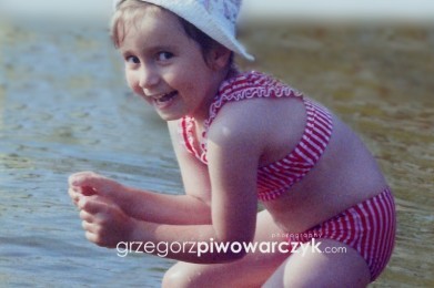 Rozbawiona dziewczynka w wieku około 5 lat stojąc w jeziorze w zabawnej pozie patrzy w obiektyw aparatu. Całość sesji utrzymana jest w klimacie retro.